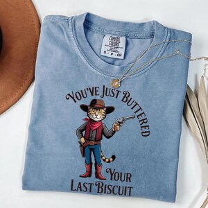 A blue t-shirt with a cartoon image of a cat holding a gun and the text "You've just BUTTERED Your LAST BISCuit" printed on it.