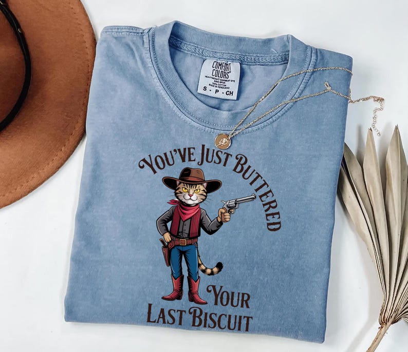 A blue t-shirt with a cartoon image of a cat holding a gun and the text "You've just BUTTERED Your LAST BISCuit" printed on it.