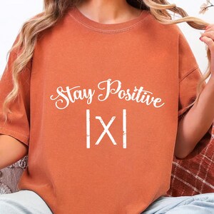 May include: Burnt orange t-shirt with the words "Stay Positive" in a white script font. Below the text is the symbol |X|. The shirt is a solid color and has short sleeves.