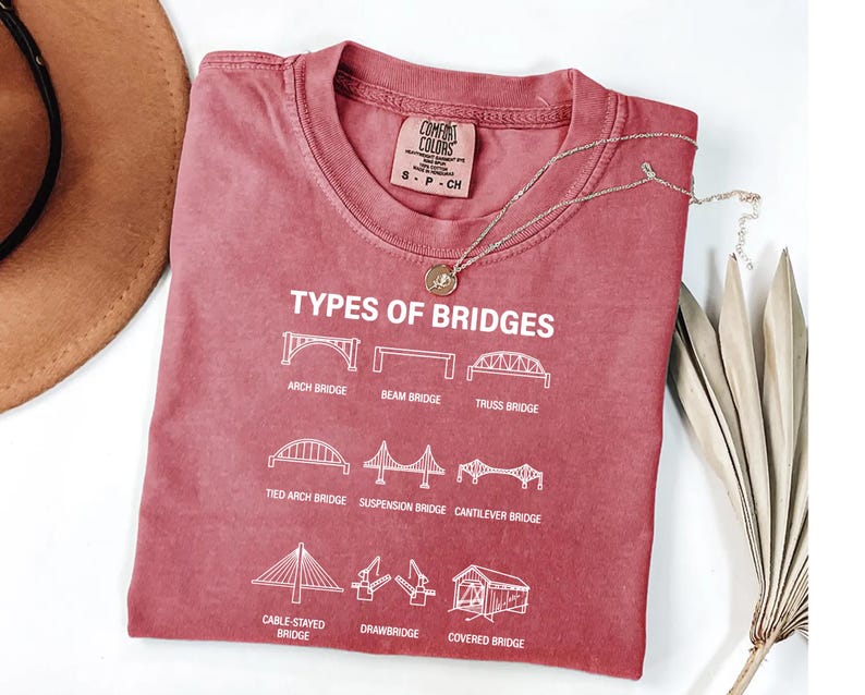A red t-shirt with white text and illustrations of different types of bridges, including an arch bridge, beam bridge, truss bridge, tied arch bridge, suspension bridge, cable-stayed bridge, and drawbridge, along with a necklace and a hat.