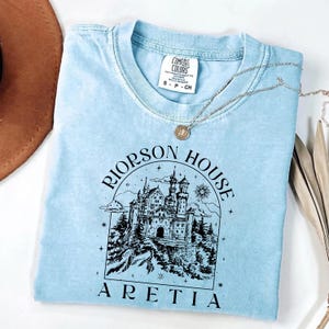 A light blue t-shirt with a graphic design of a castle and the text "Riinson House Areia" printed on it.
