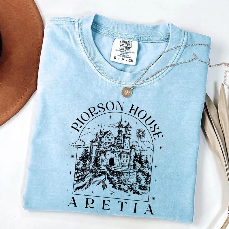 A light blue t-shirt with a graphic design of a castle and the text "Riinson House Areia" printed on it.