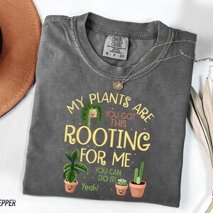 a gray t-shirt with a humorous text and illustrations of potted plants, along with a necklace and a hat.