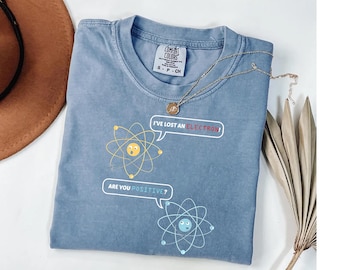 May include: A blue Comfort Colors t-shirt with a science-themed graphic. The graphic features two atom illustrations with speech bubbles that read "I've lost an electron!" and "Are you positive?" A gold necklace is draped across the shirt.