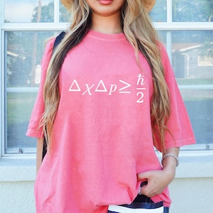 May include: A pink t-shirt with white text displaying the Heisenberg uncertainty principle formula: ΔxΔp≥ℏ/2. The shirt has short sleeves and a relaxed fit. The model is wearing a straw hat and a silver bracelet.