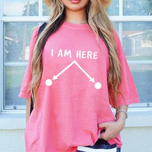 May include: A pink t-shirt with the text "I AM HERE" in white. Below the text is a white graphic with two arrows pointing outwards from a central point. The shirt has short sleeves and a crew neck.
