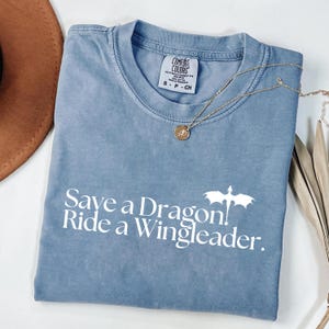 A blue t-shirt with the text "Save a Dragon. Ride a Wingleader." printed on it, along with a small dragon design. The t-shirt is folded and placed on a white surface, with a brown hat and some leaves visible in the background.