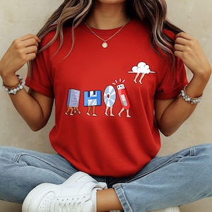 May include: A red t-shirt featuring a whimsical design of retro computer storage devices with legs, including a floppy disk, CD, and USB drive. A cloud with legs is also present. The design is in a cartoon style.