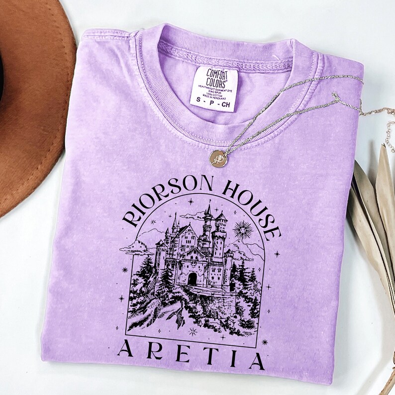 A purple t-shirt with a graphic design of a castle and the text "Rierson House Areia" printed on it, along with a necklace and a brown hat.