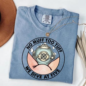 May include: A light blue Comfort Colors t-shirt with a round graphic featuring a vintage diving helmet and the text "NO MUFF TOO TUFF WE DIVE AT FIVE". A gold necklace is draped across the shirt.