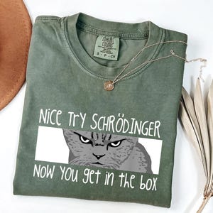 May include: Olive green t-shirt with the text "NICE TRY SCHRÖDINGER NOW YOU GET IN THE BOX" and a graphic of a grumpy cat. A gold necklace with a pendant is draped on the shirt.