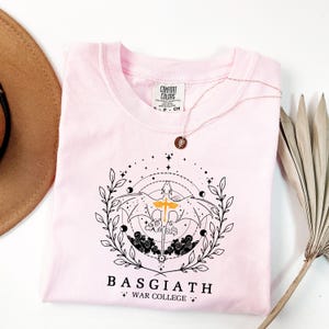 A pink t-shirt with a graphic design and text, along with a brown hat and some dried leaves, are arranged on a white background.