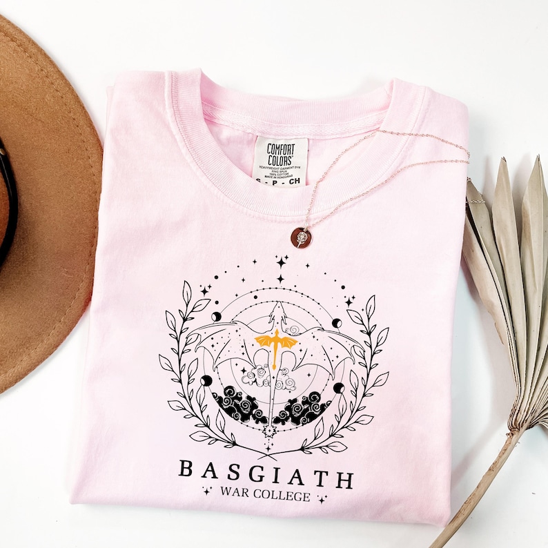 A pink t-shirt with a graphic design and text, along with a brown hat and some dried leaves, are arranged on a white background.