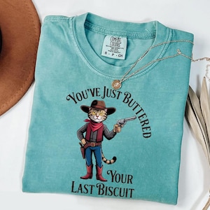 A turquoise t-shirt with a cartoon cowboy holding a gun and the text "You've just BUTTERED Your LAST BISCuit" printed on it.