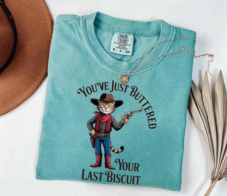 A turquoise t-shirt with a cartoon cowboy holding a gun and the text "You've just BUTTERED Your LAST BISCuit" printed on it.
