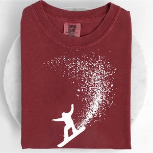 Könnte beinhalten: Ein weinrotes Comfort Colors T-Shirt mit einer weißen Grafik eines Snowboarders, der eine Kurve fährt, mit einem Spritzer weißen Schnees. Das Shirt ist ordentlich gefaltet und zeigt das Design. Das Etikett lautet Comfort Colors.