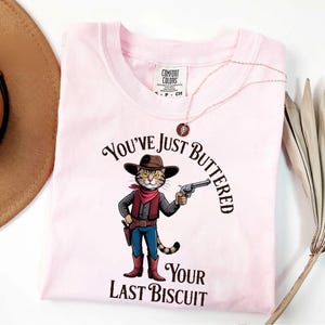 A pink t-shirt with a cartoon cat wearing a cowboy hat and holding a gun, along with the text "You've just BUTTERED Your LAST BISCUIT".
