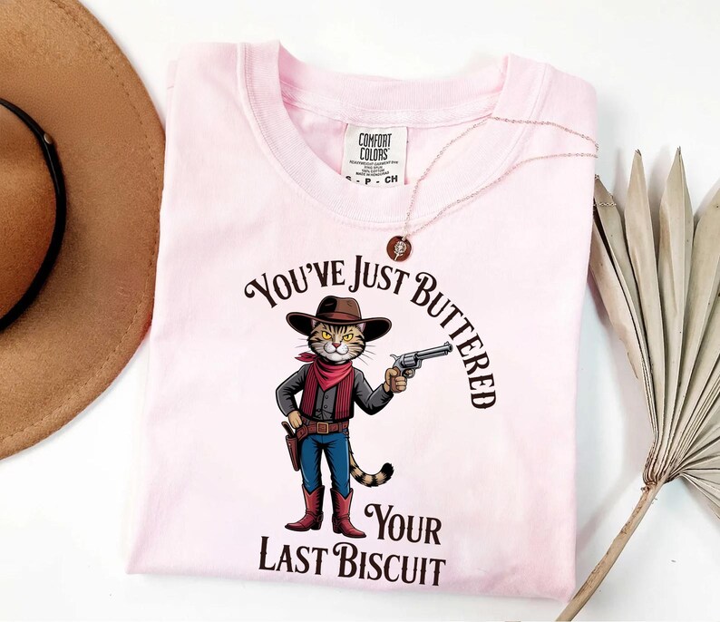 A pink t-shirt with a cartoon cat wearing a cowboy hat and holding a gun, along with the text "You've just BUTTERED Your LAST BISCUIT".