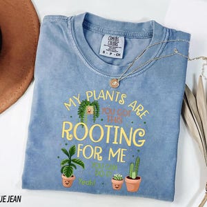 A blue t-shirt with a printed message about plants, along with a potted plant and a hat, is displayed on a white surface.