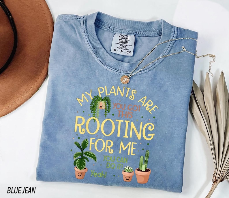 A blue t-shirt with a printed message about plants, along with a potted plant and a hat, is displayed on a white surface.