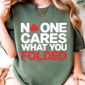 Puede incluir: Camiseta verde oliva con el texto "NO ONE CARES WHAT YOU FOLDED" en blanco y rojo. La palabra "ONE" tiene un símbolo de pica rojo encima. La camiseta es de estilo casual.