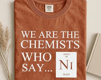 May include: Rust-colored t-shirt with the text "WE ARE THE CHEMISTS WHO SAY..." in white. A white square with "Ni" and the atomic number and weight of Nickel is printed on the shirt. The shirt is folded.