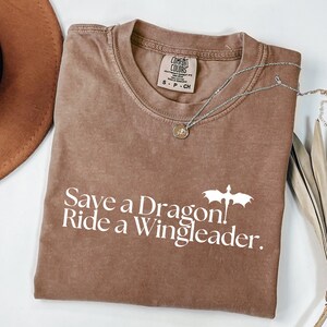 a brown t-shirt with white text that says "Save a Dragon. Ride a Wingleader." The t-shirt is folded and placed on a white surface, with a brown hat and some feathers nearby.