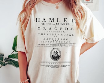May include: A cream-coloured t-shirt displaying the title page of Shakespeare's Hamlet. The text reads "Hamlet, Prince of Denmark: A Tragedy" and the author's name, William Shakespeare. The design is in black ink.