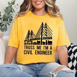 May include: Yellow short-sleeved t-shirt with a black graphic of a bridge and the text "TRUSS ME I'M A CIVIL ENGINEER". The shirt is made of a soft material and is suitable for casual wear.