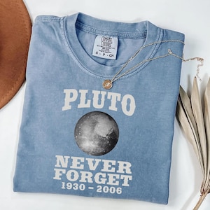May include: A light blue t-shirt with the word "PLUTO" in white block letters above a grayscale image of Pluto. Below the planet is the text "NEVER FORGET 1930-2006". A gold necklace with a circular pendant rests on the shirt.