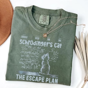 May include: Olive green t-shirt with the text "Schrödinger's cat" and a graphic of a cat. The shirt also features the text "THE ESCAPE PLAN" and the names of scientists. A gold necklace with a pendant is on the shirt.