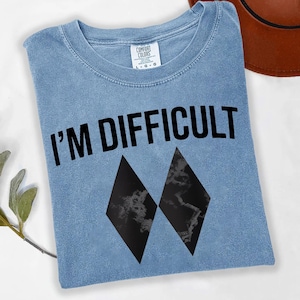 May include: Pale blue t-shirt featuring the phrase "I'M DIFFICULT" in black, bold lettering. Two black diamond shapes are below the text. The shirt has a crew neck and is made of a soft material.