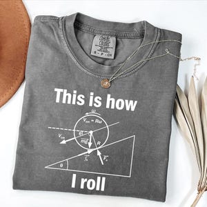 Puede incluir: Una camiseta gris Comfort Colors con el texto "This is how I roll" y un diagrama de física impreso en blanco. Un collar plateado con un colgante está sobre la camiseta. Un sombrero marrón es visible en el fondo.