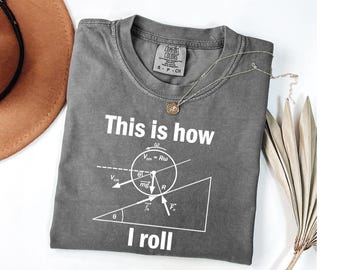 May include: A gray Comfort Colors t-shirt with the text "This is how I roll" and a physics diagram printed in white. A silver necklace with a pendant is on the shirt. A brown hat is visible in the background.