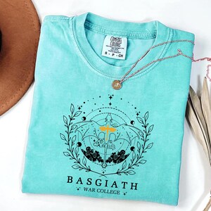 A turquoise t-shirt with a graphic design and text, along with a necklace and a brown hat, are displayed on a white surface.
