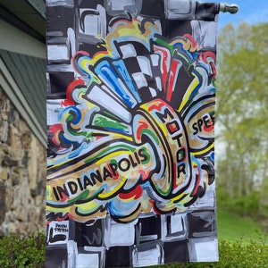May include: A colorful flag with a checkered background and a stylized design of a wheel with the words "Indianapolis Motor Speed" in a circle. The flag is hanging from a pole.