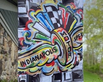 Op de afbeelding: Een kleurrijke vlag met een geruite achtergrond en een gestileerd ontwerp van een wiel met de woorden "Indianapolis Motor Speed" in een cirkel. De vlag hangt aan een paal.
