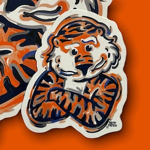 May include: A white sticker with a cartoon tiger wearing a blue and orange striped shirt. The tiger is smiling and has its arms crossed. The sticker has the text "Justin Patten" at the bottom.