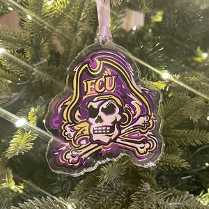 May include: Clear acrylic Christmas ornament featuring a pirate skull and crossbones design in purple, gold, and white. The ornament has the letters "ECU" in gold on the pirate hat. The ornament hangs from a silver ribbon.