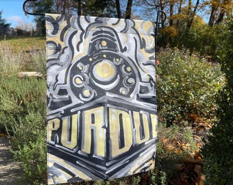 Könnte beinhalten: Eine schwarz-weiße Gartenflagge mit einer stilisierten Illustration einer Lokomotive und dem Wort "Purdue" in gelben und weißen Buchstaben.