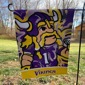 May include: A purple and yellow garden flag with a cartoon Viking wearing a purple helmet and holding a football. The flag has the word "VIKINGS" printed on the bottom.