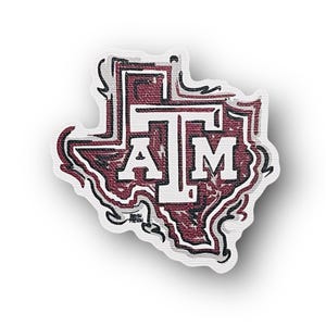 May include: A maroon and white patch in the shape of Texas, featuring the letters "ATM" in a bold, blocky font. The patch has a textured appearance with a white border and black accents.