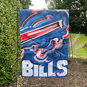 May include: A blue garden flag with a colorful abstract design featuring red, white, and blue brushstrokes. The word "BILLS" is displayed in large white letters at the bottom, with the team logo. The flag is hanging outdoors.