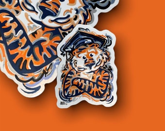May include: A collection of stickers featuring a cartoon tiger wearing a graduation cap. The tiger is depicted in orange, navy blue, and white, with a stylized design. The stickers are on an orange background.