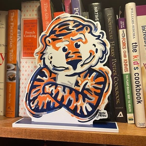 May include: A decorative cutout of a tiger mascot in orange, white, and navy blue, with arms crossed. The artwork is displayed on a shelf with cookbooks and other books. The artist's signature is visible.