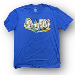 May include: A royal blue t-shirt featuring the text "Indy 500" in a colorful, stylized font. The design includes a depiction of race cars. The shirt is made of a soft material and has short sleeves.