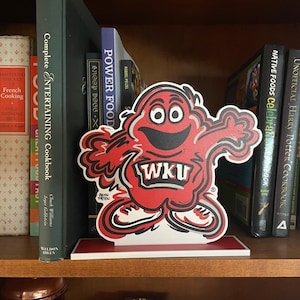 May include: A red and white cartoon mascot bookend with the letters WKU on the front. The bookend is sitting on a bookshelf with a variety of books.