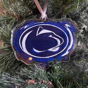 May include: A clear, oval-shaped Christmas ornament featuring the Penn State Nittany Lion logo in white and navy blue. The ornament hangs from a pink ribbon against a backdrop of green pine needles.