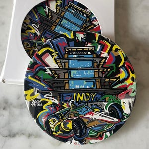 May include: Round coaster with a colorful graffiti-style design featuring a race car, the word "INDY," and a multi-story building. The coaster is in a white box, with a second coaster visible.