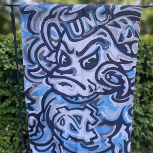 May include: A rectangular flag featuring a stylized design in shades of blue, gray, and white. The design includes the word "UNC" and a graphic of a snarling mascot. The flag is hanging outdoors against a backdrop of green foliage.